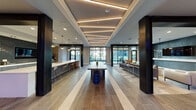 Located at 1701 Central Ave in Saint Petersburg, Florida, this contemporary amenity lounge is designed to impress with its open, single-floor layout and thoughtfully curated interior. The space is anchored by a dramatic coffered ceiling adorned with recessed linear LED lighting, casting a warm, ambient glow throughout the main corridor. Bold dark structural columns frame the open floor plan, adding an industrial-modern contrast to the light-toned large-format tile flooring, which features subtle striped inlays that guide the eye through the space. A long communal wood table serves as a central gathering point, flanked by barrel-style seating. At one end, a vivid, large-scale mural with bold sunset and ocean motifs adds an energetic focal point and nods to the Florida coastal aesthetic. Floor-to-ceiling windows and glass entry doors flood the interior with natural light and provide views of the surrounding streetscape. A built-in kitchen bar with white countertops, bar stools, and pendant lighting offers a casual dining and social hub. Adjacent lounge areas feature curated seating arrangements around a wall-mounted TV set against a textured dark accent wall. Private glass-door rooms provide quieter, enclosed spaces for meetings or focused work. The overall ambiance balances vibrant creativity with refined, modern comfort.