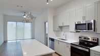 This single-floor residence showcases a clean, contemporary design throughout. The open-concept kitchen is a standout feature, outfitted with light grey shaker-style cabinetry, white quartz countertops, a white subway tile backsplash, and a full suite of stainless steel appliances including a refrigerator, range, microwave, and dishwasher. A large central island with a sleek white surface provides additional prep and dining space, while stylish dome pendant lights hang overhead, adding a polished touch.

The living area is generously sized, featuring warm wood-look flooring, a ceiling fan, and a full-width sliding glass door that floods the space with natural light and provides outdoor access. The bright white walls and open layout contribute to an airy, welcoming ambiance.

The bathroom is elongated and functional, featuring a dual-sink vanity with white quartz countertops, a framed mirror with vanity lighting, wood-look flooring, and access to a walk-in closet with built-in shelving. Additional bedrooms are carpeted, naturally lit through large windows, and equipped with ceiling fans, maintaining a consistent, comfortable aesthetic throughout the residence.