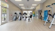 The fitness center at Springs At LaGrange offers a spacious, thoughtfully designed workout facility on a single floor. The open layout is enhanced by light wood-style flooring, neutral warm-gray walls, and abundant overhead fluorescent lighting complemented by ceiling fans, creating a clean and energizing atmosphere. Large glass garage-style doors and oversized windows flood the space with natural light while offering views of the surrounding outdoor areas, adding a refreshing sense of openness.

The cardio zone is generously equipped with multiple elliptical trainers, treadmills, and a stationary cycling bike, all positioned to take advantage of the natural light and views. Mounted flat-screen televisions are strategically placed throughout for entertainment during workouts. A dedicated strength training area features a full rack of kettlebells and dumbbells in varying weights, a weight bench, and a multi-function cable machine, supported by large wall mirrors that enhance the sense of space.

Functional training is also well-represented, with a core exercise station, resistance bands, a SmartMat with a posted exercise guide, and a collection of stability and exercise balls stored in a corner rack. The facility is thoughtfully organized to support a wide range of fitness routines, making it a comprehensive amenity for residents of Springs At LaGrange.