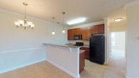 The Highrock with Sunroom is a well-appointed single-floor residence offering two bedrooms and two full bathrooms across a generously sized floor plan. The kitchen serves as a functional centerpiece, featuring rich cherry-toned cabinetry, granite countertops, and black appliances including a range, built-in microwave, dishwasher, and refrigerator. A peninsula with corbel-supported overhang provides casual seating and separates the kitchen from the adjoining dining and living areas. Pendant lighting and crown molding add refined detail throughout the main living space, which transitions seamlessly into the sunroom—a bright, windowed area with a pendant light fixture and views of the surrounding neighborhood. Both bedrooms are carpeted and offer ample natural light through large windows with blinds; one bedroom includes an attached walk-in closet. The two bathrooms are finished consistently with the kitchen, featuring granite-topped vanities, cherry cabinetry, and tile flooring. The overall aesthetic is clean and classic, with crown molding, neutral wall tones, and tile flooring in wet areas complementing the warm carpeting in living and sleeping spaces.