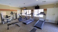 Located in Midland, TX, this single-floor fitness center is thoughtfully equipped to meet a variety of workout needs. The space features multiple treadmills and an elliptical trainer positioned along the windowed wall, allowing users to exercise while enjoying natural light that filters in through the slatted blinds. A stationary bike and a dedicated free weight area with a medicine ball rack and weight station round out the cardio and strength training options, while a flat workout bench adds further versatility. Two wall-mounted flat-screen TVs are strategically positioned throughout the room, keeping users entertained during their workouts. The interior design features warm earth-toned walls accented by a distinctive horizontal stripe of deep red and orange, adding a vibrant yet functional aesthetic to the space. Crown molding along the ceiling and recessed lighting contribute to a clean, polished finish. Carpeted flooring covers the workout area, providing cushioning underfoot. The overall layout is open and well-organized, making efficient use of the available space while maintaining a comfortable and motivating environment for exercise.
