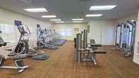 This fitness center offers a clean, functional environment designed to accommodate a range of exercise needs. The space is outfitted with a row of cardio machines, including ellipticals and treadmills, all equipped with individual display monitors for tracking workout metrics. Complementing the cardio section, a selection of strength training equipment is positioned toward the center of the room, providing options for targeted muscle conditioning. The floor is covered in durable, textured carpet in warm earth tones, offering a practical surface suited for a fitness environment. Overhead, a dropped ceiling with recessed fluorescent panel lighting ensures the space is evenly and brightly illuminated throughout. Natural light is introduced through windows along one wall, enhancing the overall brightness of the room. The walls are finished in a neutral tone, contributing to a clean and focused atmosphere. A private door is visible at the rear of the space, suggesting access to an additional room or utility area. The overall layout is open and organized, allowing for comfortable movement between equipment stations.