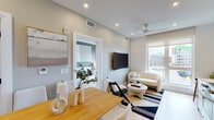 This thoughtfully designed single-floor residence seamlessly blends contemporary style with functional living. The open-concept layout connects the dining area and living room, where light wood flooring, recessed lighting, and large windows invite abundant natural light and views of the surrounding neighborhood. The living area features a wall-mounted TV, a ceiling fan, and a cozy seating arrangement anchored by a striped area rug, creating a warm and inviting atmosphere.

The dining area accommodates a natural wood table with upholstered chairs, complemented by a round mirror and curated wall art. Adjacent to it, the modern kitchen is outfitted with stainless steel appliances — including a range, microwave, and dishwasher — flat-front cabinetry in a warm wood tone, white quartz countertops, and a striking marble-look tile backsplash that adds an elegant touch.

The bedroom is a tranquil retreat, centered around a king-sized bed framed by mid-century modern nightstands and soft, layered lighting. A large window allows natural light to fill the room, while a ceiling fan adds comfort. A sleek, mirrored sliding door leads to a well-organized walk-in closet with built-in shelving and drawers.

The bathroom is a standout feature, finished floor-to-ceiling in white marble-look tile. It includes a floating wood vanity with a white countertop and a spacious glass-enclosed walk-in shower with a rain showerhead, delivering a spa-like experience.