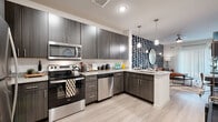 This single-floor residence presents a cohesive modern design highlighted by consistent use of dark wood-grain cabinetry, light wood-style flooring, and stainless steel accents. The kitchen serves as a central feature, outfitted with full stainless steel appliances including a range, microwave, dishwasher, and refrigerator. Granite-style countertops, a classic subway tile backsplash, and generous upper and lower cabinetry provide both functionality and visual appeal. Pendant lighting and an adjacent bold black-and-white patterned accent wall add a striking design element that extends into the living area.

The living space is bright and open, benefiting from large windows dressed with sheer curtains that invite natural light. The bedrooms continue this airy quality, each featuring wide windows, plush carpeting, and ceiling fans. One bedroom offers a notably spacious walk-in closet with dual hanging rods and built-in shelving. The bathroom mirrors the kitchen's aesthetic with dark wood-grain vanity cabinetry, granite-style countertops, a large vanity mirror, and a shower-tub combination. Light wood-style flooring flows through the main areas, tying the overall design together cohesively.