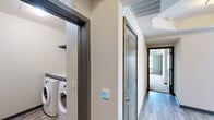 This single-floor, 2-bedroom, 3-bathroom residence offers a clean, modern aesthetic with a practical and well-organized layout. The living area features wood-look plank flooring and direct access to an outdoor balcony through a bold dark-framed glass door, providing pleasant urban views of the surrounding cityscape. The kitchen is outfitted with granite-style countertops, gray-toned cabinetry, stainless steel appliances, and matching wood-look flooring that carries through from the living space, creating a cohesive open feel. A tray ceiling detail at the kitchen-to-hallway transition adds a subtle architectural touch. The hallways connect the living spaces to the bedrooms and bathrooms with efficiency, and feature smart thermostat controls and large overhead ventilation. The spacious primary bedroom includes plush carpet flooring, a ceiling fan, and a well-sized window offering natural light and city views. Its walk-in closet is outfitted with a built-in organizational system featuring shelving towers, hanging rods, and a central shelf unit. The primary bathroom offers a glass-enclosed tile shower with stone-look tile surround and a granite-accented bench. The dedicated laundry room includes a full-size washer and dryer beneath a granite countertop with overhead shelving for added utility.