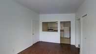This single-floor unit presents a straightforward, functional layout centered around an open living area. The living space is finished with dark hardwood flooring that contrasts with the bright white walls, creating a clean and simple aesthetic throughout. Natural light enters through the adjacent room, contributing to an airy feel despite the compact footprint.

A pass-through window connects the living area to the kitchen, providing an open sightline and a sense of connectivity between the two spaces. The kitchen is equipped with a stove and a full-size refrigerator, and features light-toned cabinetry with a tiled backsplash. The cabinetry offers ample overhead storage, while the countertop layout provides a practical workspace. The kitchen flooring transitions to a lighter tile, distinguishing it from the living area.

An adjacent room, visible through a doorway beside the kitchen, appears to benefit from natural light coming through a window, suggesting it could serve as a bedroom or additional functional space. The entry door, fitted with gold-toned hardware, opens directly into the living area, offering a straightforward and accessible entry point into the unit.