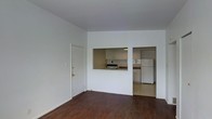 This single-floor unit presents a straightforward, functional layout centered around an open living area. The living space is finished with dark hardwood flooring that contrasts with the bright white walls, creating a clean and simple aesthetic throughout. Natural light enters through the adjacent room, contributing to an airy feel despite the compact footprint.

A pass-through window connects the living area to the kitchen, providing an open sightline and a sense of connectivity between the two spaces. The kitchen is equipped with a stove and a full-size refrigerator, and features light-toned cabinetry with a tiled backsplash. The cabinetry offers ample overhead storage, while the countertop layout provides a practical workspace. The kitchen flooring transitions to a lighter tile, distinguishing it from the living area.

An adjacent room, visible through a doorway beside the kitchen, appears to benefit from natural light coming through a window, suggesting it could serve as a bedroom or additional functional space. The entry door, fitted with gold-toned hardware, opens directly into the living area, offering a straightforward and accessible entry point into the unit.