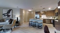This single-floor residence showcases a thoughtfully designed open-concept layout that seamlessly connects the living, dining, and kitchen areas. Light hardwood flooring runs throughout the space, lending a warm and cohesive feel to the interior. The kitchen is a standout feature, equipped with rich dark wood cabinetry, stainless steel appliances including a French door refrigerator, and a sleek white island with waterfall-style countertops. Three modern bar stools with circular bases line the island, creating an inviting casual dining area. Pendant lighting hangs above the island, complementing a track lighting system that illuminates the kitchen workspace. The adjacent living area benefits from the open plan, allowing natural flow and interaction between spaces. Neutral toned walls in soft grey and beige tones create a calm, contemporary ambiance throughout. A corridor leads to additional rooms, with interior doors finished in a consistent neutral palette. The overall design aesthetic is clean and modern, blending functional elements with stylish touches to create a comfortable and well-appointed living environment.