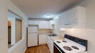 This single-floor unit offers a practical and well-organized layout designed for comfortable everyday living. The kitchen is outfitted with white cabinetry providing ample storage, light countertops, and a full suite of appliances including a coil-burner electric range, dishwasher, and refrigerator. Warm wood-look flooring runs through the kitchen, adding a welcoming contrast to the crisp white cabinetry. A pass-through window connects the kitchen to the adjacent dining or living area, promoting an open and connected feel. The living area transitions to soft carpet underfoot and features a ceiling fan with light fixture, a baseboard heating unit, and neutral wall tones that create a clean, versatile backdrop. A separate carpeted room, suitable as a bedroom, benefits from natural light through a horizontal-blind window and includes its own baseboard heater. The bathroom is finished with sage-green tile wainscoting, a combination tub and shower enclosure, and mosaic tile flooring, lending a timeless character to the space. Textured ceilings are present throughout, and the overall palette of neutral beiges and whites gives the unit a bright, move-in-ready appearance.
