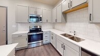 This single-floor residence offers a clean, contemporary design throughout its thoughtfully laid-out spaces. The heart of the home is an open-concept kitchen and living area, where a large white quartz island takes center stage, providing generous prep and gathering space. The kitchen is fully equipped with stainless steel appliances — including a French door refrigerator with ice maker, electric range with over-the-range microwave, and dishwasher — complemented by gray shaker-style cabinetry, a classic white subway tile backsplash, and a double-basin stainless steel sink. Pendant lighting above the island adds a stylish, functional touch. Dark wood-look flooring flows seamlessly from the kitchen into the open living area, which is brightened by a window with natural light. The bedrooms feature plush carpeting, ceiling fans, and windows that bring in additional daylight. The bathroom is finished with large-format light tile flooring, a gray vanity with white countertop, and a full soaking tub with shower. A dedicated laundry area with a full-size washer and dryer and wire shelving provides practical convenience. The overall aesthetic is modern and cohesive, with consistent use of dark trim accents, neutral tones, and quality finishes throughout.