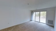 This single-floor residential space offers a clean, functional layout designed for comfortable everyday living. The main living area is a bright, open room finished with wall-to-wall carpeting and crisp white walls that create a neutral, move-in-ready canvas. A textured ceiling adds subtle visual interest overhead, while the space benefits from abundant natural light streaming in through large sliding glass doors. These doors open directly onto a private balcony enclosed by a classic railing, offering pleasant outdoor views of surrounding trees and greenery — a serene backdrop that brings a sense of nature into the interior. The sliding glass door configuration also allows for excellent ventilation and an effortless connection between the indoor living space and the outdoor deck area. The overall ambiance is light, airy, and understated, with a simple and practical design that prioritizes openness and ease of use. The neutral finishes throughout provide a versatile foundation, making the space well-suited to a variety of furnishing styles and personal tastes. Whether used as a primary residence or otherwise, this space combines everyday practicality with a calming, nature-adjacent atmosphere.