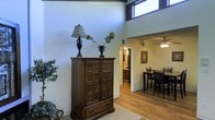 This single-story space makes a strong architectural impression through its dramatically high ceilings, which are accentuated by clerestory windows positioned near the roofline. These elevated windows allow natural light to filter into the main living area, brightening the neutral-toned walls and creating an airy, open atmosphere. The flooring throughout is warm hardwood, lending a cohesive and inviting character to the space.

The main living area serves as an entry point and transitional hub, currently styled with a substantial dark wood armoire that highlights the traditional design sensibilities present throughout. A ceiling fan is visible in the adjacent dining area, which accommodates a full dining set and connects seamlessly to the rest of the layout. The open sightlines between these areas reinforce the sense of spaciousness despite being a single level.

Beyond the dining space, a hallway leads to additional rooms, including what appears to be a bathroom or vanity area with cabinetry visible in the background. A framed artwork piece adorns the wall near the dining area, contributing to the finished, well-appointed feel of the overall space. The combination of high ceilings, natural light, and warm wood tones gives this home a welcoming and generously scaled ambiance.
