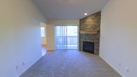 This single-floor space centers around a well-appointed living area designed for comfort and style. The focal point of the room is a striking floor-to-ceiling stacked stone fireplace, complete with a wood mantel, adding a warm and rustic character to the space. Adjacent to the fireplace, a sliding glass door fitted with vertical blinds opens to an outdoor area, allowing natural light to pour into the room while providing direct exterior access. The living area is finished with plush wall-to-wall carpeting, contributing to a cozy atmosphere underfoot. Recessed lighting in the ceiling supplements the natural light, ensuring the space remains well-lit. The smooth white walls and clean lines throughout create a neutral backdrop that complements the stone fireplace's texture. A passageway visible from the living area connects to an adjacent room with hardwood-style flooring, suggesting a varied and thoughtfully designed interior layout. Overall, the space balances functional design with inviting elements, making it a comfortable and visually appealing environment.
