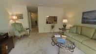 This two-floor residence offers a comfortable and well-appointed living environment with a thoughtfully designed layout. The living room serves as the heart of the home, featuring soft wall-to-wall carpeting, smooth neutral-toned walls, and ample space for relaxed seating arrangements. A distinctive architectural pass-through window connects the living area to the dining space beyond, adding an open, communicative flow between the two rooms. The dining area, partially visible through the pass-through, is illuminated by a classic chandelier that lends an understated elegance to the space. A hallway extending from the living room hints at additional rooms and functional spaces distributed across the floor. The overall ambiance is warm and inviting, with a light, airy color scheme of soft creams and beiges that carries consistently throughout. The second floor adds additional living space, expanding the overall footprint of the home. The interior design leans toward a traditional aesthetic, with decorative lighting, ornate furniture accents, and tasteful décor elements that contribute to a cozy, lived-in atmosphere. This space combines practicality with classic charm, making it a comfortable and functional multi-level home.