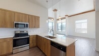 This single-floor unit showcases a thoughtfully designed open-concept layout with soaring ceilings that amplify the sense of space throughout. The kitchen is a standout feature, equipped with warm honey-toned wood cabinetry extending to the upper walls, a full suite of stainless steel appliances including a refrigerator, electric range, built-in microwave, and dishwasher. Light-toned countertops provide generous workspace, complemented by a subway tile backsplash and an undermount sink positioned on the peninsula. Pendant lighting adds an elegant finishing touch above the island.

The living area is bright and airy, featuring light hardwood-style flooring and floor-to-ceiling corner windows that frame sweeping urban skyline views. Decorative columns with wood crown detailing add architectural character to the space. The bedrooms offer plush neutral carpeting, large windows fitted with horizontal blinds, and additional city views. The bathroom is clean and functional, featuring a full soaker tub with a tiled surround, chrome fixtures, and wood-toned cabinetry. Throughout, a neutral white and warm wood palette creates a cohesive, modern aesthetic.