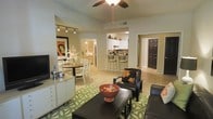 This single-floor residence showcases an open-concept design that seamlessly connects the living, dining, and kitchen areas into one cohesive and functional space. The living area serves as the central hub, featuring a ceiling fan overhead and track lighting along the wall, providing versatile illumination. Light-toned flooring runs throughout, contributing to a bright, airy atmosphere. A media console lines one wall of the living area, complemented by dark-upholstered seating arranged around a dark wood coffee table atop a green patterned area rug, creating a comfortable and stylish focal point.

Adjacent to the living space, a compact dining area accommodates a round table with white lattice-back chairs, ideal for casual meals. The kitchen, visible through a pass-through opening, features white cabinetry, a raised breakfast bar with bar seating, and ample counter space. A hallway visible from the living area leads to additional rooms, including what appears to be a bedroom accessed through a dark-paneled door. The warm, neutral wall tones throughout lend the space a welcoming and cohesive ambiance.