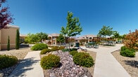 This three-story residential complex showcases a warm Southwestern architectural aesthetic, with stucco exteriors finished in earthy tan and terracotta tones, arched balconies, and pitched rooflines that blend harmoniously with the surrounding landscape. The buildings are arranged across a spacious campus-style grounds, connected by a network of wide concrete walkways equipped with metal handrails, ensuring accessible pathways throughout the community.

The outdoor common areas are thoughtfully designed for both function and relaxation. A covered pavilion with decorative stucco columns serves as a central gathering point, furnished with a swinging bench and picnic-style seating — ideal for enjoying the abundant natural sunlight. Multiple outdoor picnic tables are distributed along the main pedestrian corridor, encouraging community interaction in the open air.

The landscaping is a defining feature of the property, blending water-wise xeriscape design with lush green lawn areas. Decorative gravel, natural boulders, and rock-lined dry creek beds are interspersed with ornamental trees including vibrant red-leafed varieties, tall columnar cypress, and shade-providing deciduous trees. Manicured shrubs and seasonal flowering plants add color and texture throughout. The exterior stairways connect each floor with easy access, while the generous open sky and desert-influenced surroundings lend the entire complex a bright, airy, and welcoming ambiance.