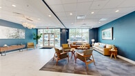 Located at 921 Manatee Ave W in Bradenton, Florida, this thoughtfully designed single-floor common area serves as a welcoming and functional hub. The space is anchored by a sophisticated lobby lounge featuring bold navy and teal accent walls that contrast beautifully with a pristine marble-look tile floor running throughout. Large floor-to-ceiling windows at the main entrance flood the space with natural light and offer views of the surrounding greenery outside.

The lounge area is furnished with a curated mid-century modern aesthetic, including wood-framed accent chairs, plush sofas in warm neutral tones, and coordinating area rugs that define the seating zones. Overhead, circular pendant lights and recessed ceiling fixtures provide layered illumination beneath a coffered tile ceiling.

A staffed reception counter finished in crisp white cabinetry serves as a central service point, while framed artwork and coastal-inspired paintings add character throughout. Adjacent areas include a well-appointed mail and package room equipped with individual mailboxes, smart package lockers, and elevator access. A secondary corridor features a wall-mounted display screen and bench seating, offering a quiet transitional space. Decorative moss wall accents and potted plants bring natural elements indoors, completing the polished, welcoming atmosphere of this well-appointed space.