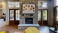 Located at 6837 N Trenholm Rd in Arcadia Lakes, SC, this single-floor community space combines elegant design with welcoming functionality. The interior is defined by dramatically high ceilings accented with board-and-batten paneling painted in a cool gray tone, lending a refined, contemporary farmhouse character throughout.

The expansive main lounge area serves as the heart of the space, anchored by a striking floor-to-ceiling stone fireplace flanked by large glass-paned doors that invite abundant natural light and outdoor views. Rich hardwood flooring flows seamlessly throughout, complementing the warm, layered furnishings that include plush leather sofas, patterned accent chairs, and decorative side tables.

Multiple seating arrangements create distinct gathering zones — one centered around a media console with open metal shelving, and another oriented toward the fireplace — offering both casual and social settings. A woven rattan mirror and curated décor accents add texture and visual interest to the walls.

The kitchen area features dark wood cabinetry, mosaic tile backsplash, stainless steel appliances, and a generously sized granite-topped bar with seating — ideal for entertaining. A globe chandelier overhead adds a stylish focal point. Large windows throughout the space frame views of the surrounding landscape, enhancing the bright, airy ambiance of this thoughtfully designed gathering venue.