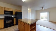 Located at 8200 W 20th St in Greeley, Colorado, this single-floor unit offers a well-designed, open layout that balances functionality and comfort. The kitchen is a central highlight, featuring abundant warm oak cabinetry, dark countertops, and a full suite of black appliances including a range, built-in microwave, dishwasher, and refrigerator. A freestanding kitchen island with open shelving below provides additional prep space and storage, while wood-look flooring throughout the kitchen and living areas adds warmth and durability. The living area flows naturally from the kitchen and is equipped with a ceiling fan, and a glass door opens to an outdoor balcony, bringing in natural light and views of the surrounding area. The bedrooms feature plush wall-to-wall carpeting and clean white walls, creating a calm and neutral environment. The bathroom is outfitted with a dual-sink vanity set in oak cabinetry with dark countertops, a large mirror, and tile flooring, offering both practicality and ample counter space. Overall, the unit presents a cohesive, functional design with consistent warm-toned wood finishes throughout, making it a comfortable and practical living space in Greeley, CO.
