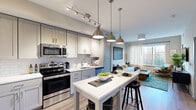 This thoughtfully designed one-bedroom, one-bathroom residence offers a smart and efficient layout across a single floor. The open-concept kitchen and living area serves as the heart of the home, where a fully equipped kitchen impresses with two-tone shaker cabinetry, white quartz countertops, a classic subway tile backsplash, and a full suite of stainless steel appliances including a refrigerator, range, microwave, and dishwasher. A central island with pendant lighting provides additional prep space and casual seating, seamlessly connecting to the living area. The living room is bright and welcoming, featuring large windows that frame outdoor views and fill the space with natural light, complemented by warm wood-look flooring that runs throughout. The bedroom is a comfortable retreat with a generously sized window overlooking a wooded outdoor scene, providing a tranquil atmosphere. A dedicated closet offers convenient storage. The bathroom features a vanity with white countertops, a deep-set sink, blue cabinetry, and a tub-shower combination with tile surround. Clean neutral tones, modern fixtures, and cohesive finishes give this residence a polished, contemporary feel from room to room.