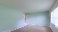 This two-floor residential space offers a clean and functional layout designed for comfortable everyday living. The main level showcases a spacious open living area finished with soft, light sage-green walls that create a calm and welcoming ambiance. Plush wall-to-wall carpeting in a neutral taupe tone runs throughout the floor, adding warmth underfoot and contributing to the overall understated aesthetic. The smooth, white ceiling complements the light wall color, giving the room an airy and open feel. A staircase with a classic black metal railing serves as the central architectural connector between the two levels, adding a subtle design accent to the otherwise minimalist space. The staircase is neatly tucked into the corner of the living area, allowing for an efficient use of floor space while maintaining an open feel in the main room. The second floor, accessible via the staircase, expands the overall living footprint of the home. Throughout the space, clean white baseboards frame each room, providing crisp definition between the walls and flooring. The overall design is neutral and move-in ready, offering a versatile canvas that can be easily personalized to a variety of interior styles.