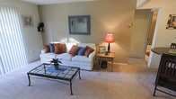 This single-floor residence offers a comfortable and thoughtfully arranged living space designed with everyday functionality in mind. The living room serves as the heart of the home, featuring wall-to-wall plush carpeting that adds warmth underfoot and contributes to the room's relaxed, cozy ambiance. Neutral, taupe-toned walls provide a versatile backdrop that complements the furnishings throughout. A generously sized white sofa anchors the seating area, accented with rust-orange and navy blue throw pillows that introduce a tasteful pop of color. A glass-top coffee table with a wrought iron base adds a touch of classic style to the space. Adjacent to the sofa, a warm-toned wooden side table holds a traditional lamp with a rich red shade, casting a soft, ambient glow. A decorative blue-framed mirror is mounted on the wall, enhancing the sense of depth and light in the room. Natural light filters into the space, further brightening the interior. The overall aesthetic is understated and welcoming, offering a move-in ready environment that balances comfort with a timeless, livable design sensibility.
