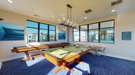 This single-floor game room in Port Wentworth, Georgia, is designed as a well-appointed recreational space that balances function with contemporary style. Large, dark-framed windows line multiple walls, flooding the interior with abundant natural light and providing views of the surrounding community and landscaping. The open layout comfortably accommodates a full-size pool table at its center, positioned beneath a modern candelabra-style pendant chandelier that serves as a striking focal point overhead. Recessed lighting complements the chandelier, ensuring consistent illumination throughout the space.

A shuffleboard table runs along one wall, adding to the variety of recreational options available. The flooring features a bold, wave-patterned deep blue carpet that adds visual interest and reinforces the room's coastal-inspired aesthetic. A live-edge wooden console table with cabinetry near the entry provides both storage and a warm, natural accent. Small bistro-style tables with teak and chrome chairs are arranged along the windowed walls, offering casual seating for spectators or socializing. Blue-toned aquatic artwork adorns the walls, tying the design theme together cohesively. The overall ambiance is energetic yet refined, making it an inviting setting for leisure and recreation.