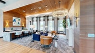 This thoughtfully designed single-floor amenity space offers a generous and versatile layout spread across two distinct areas. The main lounge is an architectural showpiece, highlighted by a warm wood-paneled ceiling, rich hardwood flooring, and floor-to-ceiling windows draped in sheer linen curtains that flood the interior with natural light and provide views of the surrounding outdoor terrace. A striking tiered crystal chandelier serves as the room's centerpiece, casting an elegant glow over the carefully curated seating arrangement, which includes a bold blue velvet sofa, cognac leather chairs, and a patterned area rug. A marble-topped bar counter along one wall, accented by a textured stone backdrop and art display, offers a refined workspace or social gathering spot. Adjacent seating nooks with linen armchairs and marble side tables provide quieter retreats beside the tall windows. The second area functions as an entertainment and gaming room, featuring a golf simulator setup, a wall-mounted television, and a lounge sofa framed by a dramatic geometric wooden screen partition. Together, these two zones create a dynamic, upscale communal environment ideal for both relaxation and recreation.