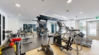 Located at 965 Av St-Charles in Saint-Lambert, Quebec, this fitness center offers a thoughtfully designed workout space across a single floor. The room makes effective use of its footprint, with distinct zones dedicated to cardio training and strength work.

The flooring transitions between warm herringbone-patterned hardwood and large-format dark rubber tiles, delineating activity areas while adding visual interest. A bold navy accent wall commands attention, displaying motivational French typography — featuring words such as "Énergie," "Défi," "Force," and "Bien-Être" — reinforcing the space's energetic atmosphere.

The cardio zone is outfitted with treadmills, elliptical trainers, and stationary bikes, including Matrix-branded equipment, all positioned to benefit from natural light streaming through full-height windows and glass doors. The strength and free-weight area features a multi-tiered dumbbell rack, resistance bands, a stability ball, a workout bench, and a cable machine, providing a comprehensive range of training options.

The ceiling features a combination of recessed pot lights and linear LED strip lighting set within a tiered tray design, creating a bright and modern ambiance. A wall-mounted air conditioning unit ensures year-round climate comfort. Large mirrors visually expand the space and support proper form during exercise. The overall design is clean, modern, and purposefully functional.