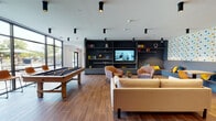 Located at 27W555 High Lake Rd in Winfield, Illinois, this single-floor community amenity space at Winfield Station is thoughtfully designed to offer residents a dynamic and inviting environment for socializing and recreation. The open-concept layout flows seamlessly across rich wood-look tile flooring, connecting multiple functional zones within one cohesive space.

The game and lounge area serves as a central hub, featuring a full-size billiards table and a shuffleboard table alongside comfortable seating arrangements. A floor-to-ceiling built-in dark shelving unit houses a mounted flat-screen TV and decorative accessories, anchored by a sleek linear fireplace below. Plush sofas and accent chairs are arranged to encourage conversation and relaxed gatherings.

A vibrant nook with a geometric-patterned feature wall offers built-in bench seating with colorful cushions and small café-style tables, providing an energetic contrast to the more subdued lounge areas.

The kitchen bar area is a standout feature, highlighted by a long wood-paneled island with bar seating, warm pendant lighting, exposed wood beam detailing on the ceiling, and a white subway tile backsplash bearing the "Winfield Station" signage.

Floor-to-ceiling windows along one wall flood the space with natural light and offer direct views of the outdoor patio area, complete with seating, grilling stations, and pergola structures, blurring the line between indoor comfort and outdoor living.