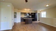 This single-floor space showcases an open-concept layout that seamlessly connects the kitchen and living areas, creating a cohesive and functional environment. The kitchen serves as the centerpiece, featuring dark espresso cabinetry that contrasts elegantly with light-toned walls. A center island with a granite-style countertop provides additional workspace and doubles as a casual dining or gathering spot. Black appliances, including a refrigerator, range, and built-in microwave, lend a sleek, uniform aesthetic to the kitchen. A stylish multi-bulb pendant chandelier hangs above the island, offering focused task lighting, while additional pendant lights and a ceiling fan with an integrated light fixture illuminate the broader living space. The warm hardwood-style flooring flows continuously throughout, enhancing the sense of openness and cohesion. A dedicated laundry area, tucked behind white cabinetry near the kitchen, adds practical convenience. Natural light filters in through windows in the adjacent living area, brightening the space and creating an inviting ambiance. The overall design balances modern finishes with a comfortable, livable feel.