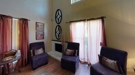 This single-story professional space is thoughtfully arranged to accommodate both client comfort and staff operations. The reception and waiting area immediately sets a welcoming tone, boasting dramatically high ceilings that lend an open, airy feel. Sheer pink curtains filter natural light from large windows, while hardwood-style flooring runs throughout. A corner fireplace serves as a focal point, flanked by plush upholstered armchairs and decorative wall art, creating a relaxed, inviting atmosphere.

Adjacent to the lounge, a small refreshment station offers a coffee maker and supplies for visitors. A glass-top table with chairs serves as a client intake area, stocked with informational brochures and paperwork. A pass-through window connects this zone to a compact back-office area, where staff can manage administrative tasks efficiently.

The administrative workspace is well-equipped with a desk, computer workstation, printer, and phone system. Framed credentials and certificates are prominently displayed on the walls, reinforcing a professional environment. Overall, the space balances a warm, hospitable ambiance in client-facing areas with functional, organized workspaces for staff.