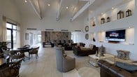 This clubhouse presents an impressive single-story layout distinguished by its dramatic vaulted ceiling with exposed white beams that draw the eye upward, creating an expansive and airy atmosphere. The open-plan lounge area is generously proportioned, offering multiple seating arrangements that cater to both intimate conversations and larger social gatherings. Dark leather sofas and plush upholstered armchairs are thoughtfully arranged throughout the space, complemented by contemporary coffee tables with gold and glass accents. The flooring consists of large, light-toned tiles that enhance the sense of openness. Built-in wall niches display decorative objects and lanterns, adding character to the crisp white walls. A striking stone or marble feature wall serves as a focal point at one end of the room, providing a bold textural contrast. Recessed spotlights and natural light from surrounding windows ensure the space is well-lit throughout the day. The overall ambiance is refined yet welcoming, blending contemporary design with warm, layered décor to create a versatile and sophisticated communal environment.