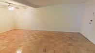 This single-floor space presents a clean and straightforward layout that offers a blank canvas for a variety of uses. The most distinctive design element is the classic parquet wood flooring, which features a traditional block pattern in warm, light-toned wood. This timeless flooring choice adds a sense of character and craftsmanship to the interior, providing both visual interest and durability underfoot. The walls are finished in a neutral, off-white tone that reflects light evenly throughout the space, contributing to a bright and open atmosphere. A subtle architectural detail is visible in the form of a shallow overhead soffit near the ceiling line, which adds a gentle sense of definition to the room without disrupting the overall openness. The smooth, uninterrupted wall surfaces provide ample flexibility for furniture arrangement and personalization. The space maintains a minimalist aesthetic, allowing the quality of the materials — particularly the parquet flooring — to serve as the central focal point. Overall, the interior conveys a calm, understated ambiance that is well-suited for a range of functional configurations.