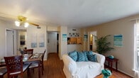 Located at 7350 McArdle Rd in Corpus Christi, Texas, this single-floor residence offers a well-organized layout with distinct living, dining, kitchen, and sleeping areas. The living room serves as the heart of the home, featuring rich hardwood-style flooring, a classic fireplace with a white mantel, and a large window dressed with vertical blinds that fills the room with natural light and offers outdoor views. The open sightlines connect seamlessly to the dining area, which includes a ceiling fan with a light fixture and space for a full dining table set. The kitchen is equipped with dark cabinetry, speckled countertops, and a double stainless steel sink, with a pass-through window that maintains visual connection to the living space. The residence includes two bedrooms, each with plush carpeting, ceiling fans, and ample natural light from well-sized windows. One bedroom features a textured ceiling and dark curtains for enhanced privacy, while the other is finished with a similar cozy aesthetic. Two bathrooms serve the home, each with shower-tub combinations and dark-toned vanity counters. Throughout, the warm, neutral color palette and consistent coastal-inspired accents give the home a cohesive and inviting character.