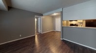 This single-floor residence offers a functional and cohesive layout finished in a consistent gray color palette throughout. The open living area is finished with dark hardwood-style flooring that extends through the main living spaces, providing a warm and unified aesthetic. A textured ceiling adds character to each room.

The kitchen is designed in a galley configuration, featuring white cabinetry with ample storage, granite-style countertops, and black appliances including a refrigerator, gas range, and dishwasher. A stainless steel sink sits beneath the counter on one side. The kitchen connects to the living area via a pass-through counter with overhead white cabinets, creating an open and communicative flow between spaces.

The living area is generously sized with a window that brings in natural light, white baseboards, and a door leading to additional rooms. A closet with white bi-fold doors provides convenient storage near the entryway.

Both bedrooms are finished with plush carpeting and gray walls, each featuring windows with blinds that allow natural light. One bedroom includes access to a closet with built-in shelving.

The bathroom features a full-size soaking tub and shower combination with white surround panels, a toilet, and a towel bar, all finished in the same gray tones found throughout the unit.