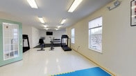 Located at 232 W. Gilbert St. in Hampton, Virginia, this single-floor fitness facility offers a clean, well-lit environment designed to support a variety of workout routines. The space is bright and open, with large windows along one wall that invite natural light and provide views of the surrounding area, creating an airy and energizing atmosphere.

The workout area is equipped with a range of cardio machines, including a treadmill and stationary bikes, making it suitable for both low-impact and high-intensity cardiovascular training. A dumbbell rack with a selection of free weights is positioned along the back wall, supporting strength and resistance training. A wall-mounted flat-screen television is installed near the equipment area, allowing users to stay entertained or informed during their workouts.

Overhead, a series of recessed fluorescent light panels ensure consistent, even illumination throughout the room, complementing the natural light from the windows. The flooring is a smooth, light-colored surface with interlocking foam exercise mats laid out in a dedicated area, ideal for stretching, floor exercises, or martial arts-style training.

The overall ambiance is clean, functional, and welcoming, with neutral white walls that keep the focus on the equipment and open floor space. This facility provides a practical and accessible environment for a well-rounded fitness routine.