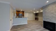 This single-floor space showcases a thoughtfully designed open-concept layout that seamlessly connects the kitchen and living area, creating a cohesive and functional environment. The kitchen is equipped with rich, warm-toned wood cabinetry, granite countertops, and stainless steel appliances, including a built-in microwave. A peninsula with a granite overhang serves as both a prep surface and a casual dining or gathering spot, effectively defining the kitchen zone while maintaining visual openness to the rest of the space. A mosaic tile backsplash adds a refined decorative touch behind the cooking area. Glass block windows near the entry allow natural light to filter in while maintaining privacy. The open living area is expansive and bright, featuring light-toned wood-look flooring that runs throughout, creating a clean and modern aesthetic. Recessed lighting is strategically placed across the ceiling to ensure even, well-distributed illumination. Crown molding adds an elegant architectural detail along the ceiling perimeter. To one side, a whitewashed brick fireplace serves as a striking focal point, complemented by built-in shelving and a built-in desk or bar area with additional cabinetry. The neutral gray wall tones and crisp white trim tie the entire space together, resulting in a polished and inviting atmosphere.