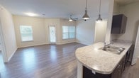 This single-story, 3-bedroom, 2-bathroom residence features a thoughtfully designed open-concept layout that seamlessly connects the living area and kitchen. Wood-style plank flooring runs throughout the entire unit, providing a cohesive and contemporary aesthetic. The spacious living area is filled with natural light from multiple windows and a glass door that leads to the exterior, complemented by a ceiling fan overhead.

The kitchen is well-appointed with dark espresso cabinetry, light granite-style countertops, black appliances including a refrigerator, electric range, and microwave, plus a curved peninsula with an undermount sink and pendant lighting above. A dedicated utility closet is accessible directly from the kitchen. Track lighting provides additional illumination over the cooking area.

Each of the three bedrooms features wood-style flooring, a ceiling fan with light fixture, and a window offering natural light and outdoor views. The two bathrooms maintain consistent finishes, featuring dark vanity cabinetry, granite-style countertops, and tub-shower combinations with white tile surrounds. The overall color palette of warm neutrals and dark accents gives the space a clean, modern character throughout.
