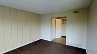 This single-story space features a simply appointed room finished with dark wall-to-wall carpeting that contrasts with the neutral, light-toned walls. One wall showcases vertical wood panel detailing, adding a subtle textural element to the otherwise understated interior. A doorway leads directly into a walk-in closet, which is equipped with built-in shelving and a hanging rod, providing practical storage capacity. The closet appears to transition into an adjoining area with lighter flooring, suggesting a connected room or hallway beyond. Overhead, a flush white ceiling is paired with a standard HVAC vent, keeping the look clean and unobtrusive. Additional wall outlets and a light switch are positioned for convenience throughout the room. The overall ambiance is minimal and functional, presenting a neutral canvas well-suited for personalization. Natural light does not appear prominently in this view, giving the space a quiet, enclosed feel. The straightforward design and built-in storage make this room particularly practical for use as a bedroom or private living area.