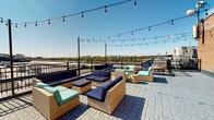 This single-floor space includes two notable features: a rooftop terrace and a half bathroom. The rooftop terrace is a standout amenity, finished with a paver tile deck and enclosed by a sturdy wrought iron railing. String lights are strung overhead between poles, creating an inviting ambiance suited for evening gatherings. The seating area is furnished with wicker sectional pieces accented in navy blue and teal cushions, arranged to take full advantage of the panoramic views of the surrounding cityscape and tree canopy stretching to the horizon. The brick exterior wall adds an urban, industrial character to the space.

The half bathroom is compact and straightforwardly appointed. Warm, neutral yellow-toned walls create a soft atmosphere. A white vanity with a wood cabinet base supports an integrated sink basin, paired with chrome fixtures and a wood-framed rectangular mirror above. A white toilet is positioned opposite, with a small decorative shelf mounted on the wall above it. White panel doors with brass hardware provide access to the space. Together, these elements present a clean, functional interior that complements the building's overall character.