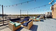 This single-floor space includes two notable features: a rooftop terrace and a half bathroom. The rooftop terrace is a standout amenity, finished with a paver tile deck and enclosed by a sturdy wrought iron railing. String lights are strung overhead between poles, creating an inviting ambiance suited for evening gatherings. The seating area is furnished with wicker sectional pieces accented in navy blue and teal cushions, arranged to take full advantage of the panoramic views of the surrounding cityscape and tree canopy stretching to the horizon. The brick exterior wall adds an urban, industrial character to the space.

The half bathroom is compact and straightforwardly appointed. Warm, neutral yellow-toned walls create a soft atmosphere. A white vanity with a wood cabinet base supports an integrated sink basin, paired with chrome fixtures and a wood-framed rectangular mirror above. A white toilet is positioned opposite, with a small decorative shelf mounted on the wall above it. White panel doors with brass hardware provide access to the space. Together, these elements present a clean, functional interior that complements the building's overall character.