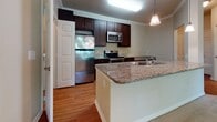 This single-floor residence located in Charlottesville, Virginia, offers a well-designed open-concept layout that seamlessly connects the kitchen, dining, and living areas. The kitchen is a standout feature, equipped with rich dark-wood cabinetry, granite countertops, and a full suite of stainless steel appliances including a refrigerator, range, and built-in microwave. A peninsula with an integrated sink provides additional prep space and serves as a natural divider between the kitchen and the living area. Warm hardwood-style flooring runs throughout the kitchen, transitioning to plush neutral carpeting in the expansive living room. Crown molding and a ceiling fan with a light fixture add refined detail to the living space, which also benefits from a sliding glass door that brings in natural light. Track and pendant lighting complement the open layout throughout. The bedroom is generously sized, carpeted, and features a window with a lush green outdoor view. The bathroom includes a dark-wood vanity with a light stone countertop, a large mirror, and tiled shower surround. Overall, the space presents a clean, functional, and comfortable living environment.