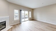 This third-floor unit in Redmond, WA offers a well-designed two-bedroom, two-bathroom layout finished with contemporary materials throughout. The open-concept living and kitchen area serves as the heart of the space, where light wood-tone luxury vinyl plank flooring extends underfoot and neutral warm-gray walls create a cohesive backdrop. The kitchen is appointed with white shaker cabinetry, quartz countertops featuring a curved peninsula with an undermount sink, stainless steel appliances, and a distinctive hexagonal tile backsplash. Pendant lighting above the peninsula adds a polished touch. The living room features a tiled fireplace and expansive sliding glass doors flanked by windows that bring in generous natural light and open to a private balcony with views of the surrounding Redmond skyline and greenery. The primary bedroom includes hardwood-style flooring and a sliding barn door that leads to a closet. The second bedroom also features the same flooring and ample wall space. Both bathrooms are finished with wood-look tile flooring, white vanities with quartz counters, and brushed nickel fixtures, with one featuring a full tub-shower combination.