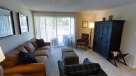 This single-floor space offers a warm and inviting atmosphere defined by classic furnishings and thoughtful design. The living room serves as the centerpiece, featuring plush carpeted flooring and a textured ceiling that adds character to the space. Large sliding glass doors lined with vertical blinds flood the room with natural light and provide direct access to an outdoor balcony or patio area, offering pleasant views of surrounding greenery. The living room is well-appointed with a comfortable seating arrangement, including a tan sofa accented with dark throw pillows, a plaid armchair, and an upholstered ottoman, creating a cozy and functional gathering area. A stately dark wood armoire anchors one corner of the room, adding a touch of traditional elegance. A classic floor lamp provides warm ambient lighting, complementing the natural light from the expansive glass doors. A framed artwork piece adorns the wall, contributing to the room's curated, homey feel. The overall ambiance of the space is relaxed and comfortable, blending traditional design elements with practical living features to create a functional and welcoming environment.