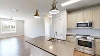 This well-appointed single-story, 4-bedroom, 2-bathroom home offers a thoughtfully designed layout that balances comfort and modern style. The open-concept kitchen serves as a centerpiece of the living area, showcasing a large island with a deep single-basin sink, quartz countertops, light wood cabinetry, subway tile backsplash, and a full suite of stainless steel appliances including a refrigerator, range, and built-in microwave. Stylish brass and nickel pendant lights illuminate the space with warm character. The kitchen flows seamlessly into the living room, which features warm luxury vinyl plank flooring, a ceiling fan, and a window offering outdoor views of lush greenery. The four bedrooms are well-sized, with the primary bedroom providing generous proportions and a walk-in closet equipped with wire shelving. Bedrooms are finished with plush carpet and windows that bring in natural light and outdoor views. The bathrooms are cleanly designed, featuring wood-toned vanities with dark countertops, subway-tiled tub-shower combinations, and wood-look flooring. Neutral gray tones, clean white trim, and consistent finishes throughout give the home a cohesive, modern aesthetic.