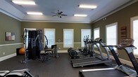 This fitness center offers a dedicated single-floor space designed for a comprehensive workout experience. The room features rich olive-green walls accented by crisp white crown molding and baseboards, lending a clean and polished aesthetic. Multiple windows fitted with white blinds line the walls, allowing natural light to fill the space while maintaining privacy. A ceiling fan with integrated lighting is centrally mounted, ensuring comfortable air circulation during workouts.

The flooring consists of durable rubber gym matting, ideal for both safety and equipment stability. The space is well-appointed with a variety of exercise equipment, including treadmills, elliptical cross-trainers, and a multi-station strength training machine, providing users with options for both cardiovascular and resistance training. Overhead, fluorescent strip lighting supplements the natural light, keeping the room consistently bright throughout the day.

The layout is thoughtfully arranged to allow ample room for movement between machines, supporting a safe and functional workout environment. The overall ambiance is practical and motivating, combining clean architectural details with purpose-driven amenities to create an inviting and fully functional fitness facility.