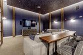 This single-level home theater is designed to deliver an immersive entertainment experience through a carefully curated combination of materials, lighting, and furnishings. The room is defined by deep navy-painted walls accented by vertical rustic wood columns that add warmth and texture against the dark backdrop. A dramatically dark textured ceiling is lined with indirect LED cove lighting, creating a soft, ambient glow that reinforces the cinematic atmosphere without harsh overhead illumination. Wall-mounted sconces provide additional layered lighting throughout the space.

The main viewing area is anchored by a large flat-screen television mounted on the feature wall above a dark carved-wood media credenza. In front of the screen, a generously sized neutral-toned sectional sofa centers the room, complemented by a striking live-edge wood coffee table that adds an organic, artisan element. Plush carpeting underfoot enhances the acoustic quality and overall comfort of the space.

Along one side of the room, a live-edge wood bar counter accompanied by upholstered bar-height chairs provides an elevated seating option for viewing or entertaining. Decorative wall art adds visual interest nearby. Additional utility is offered through a built-in countertop and cabinetry area visible toward the rear, providing convenient storage and serving space within easy reach of all seating areas.