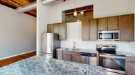 Located at 1151 Falls Rd in Rocky Mount, NC, this single-floor unit masterfully blends industrial character with modern finishes. The open living area makes an immediate impression with its rich, original hardwood floors, soaring wood-plank ceilings with exposed timber beams, and industrial ductwork that nods to the building's heritage. A dramatic wall of large, steel-framed factory windows floods the space with natural light and offers lush outdoor views, reinforcing the unit's converted-industrial aesthetic.

The kitchen is well-appointed with custom wood cabinetry, striking granite countertops with bold veining, a full suite of stainless steel appliances including a refrigerator, electric range, built-in microwave, and dishwasher, complemented by a subway tile backsplash and pendant lighting overhead. A generous granite-topped island or counter provides additional workspace and visual appeal.

The bathroom continues the space's character with original hardwood flooring, a white vanity with a marble-style countertop, a walk-in shower, and clean, modern fixtures. Throughout the unit, exposed structural columns add an authentic industrial touch. The overall ambiance is one of thoughtful adaptive reuse — combining the rawness of historic architecture with carefully selected contemporary finishes.